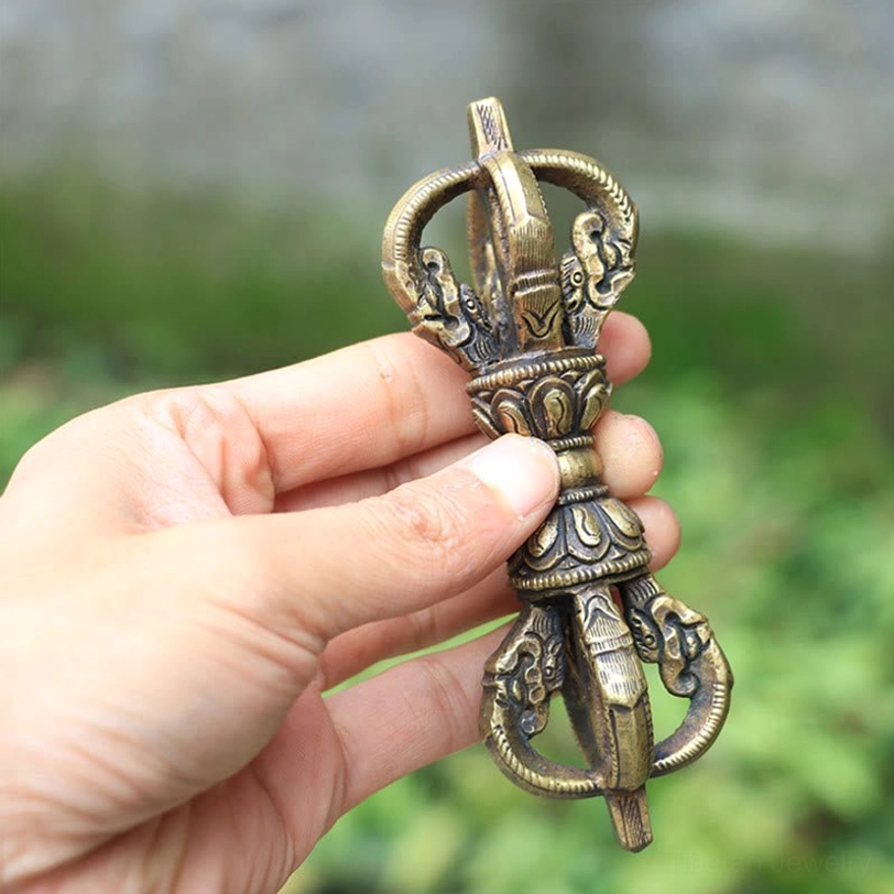 Single five-prong tibetan vajra dorje in hand held view, outdoor natural green backdrop, showing detailed ritual carvings and antique bronze metal finish with authentic aged patina