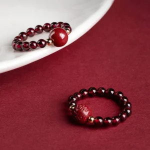 Garnet cinnabar PiXiu ring with Om Mani Padme Hum mantra engraving, displayed on white porcelain with gold metal spacer beads on wine red background