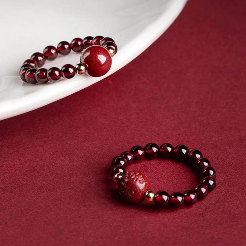 Garnet cinnabar PiXiu ring with Om Mani Padme Hum mantra engraving, displayed on white porcelain with gold metal spacer beads on wine red background