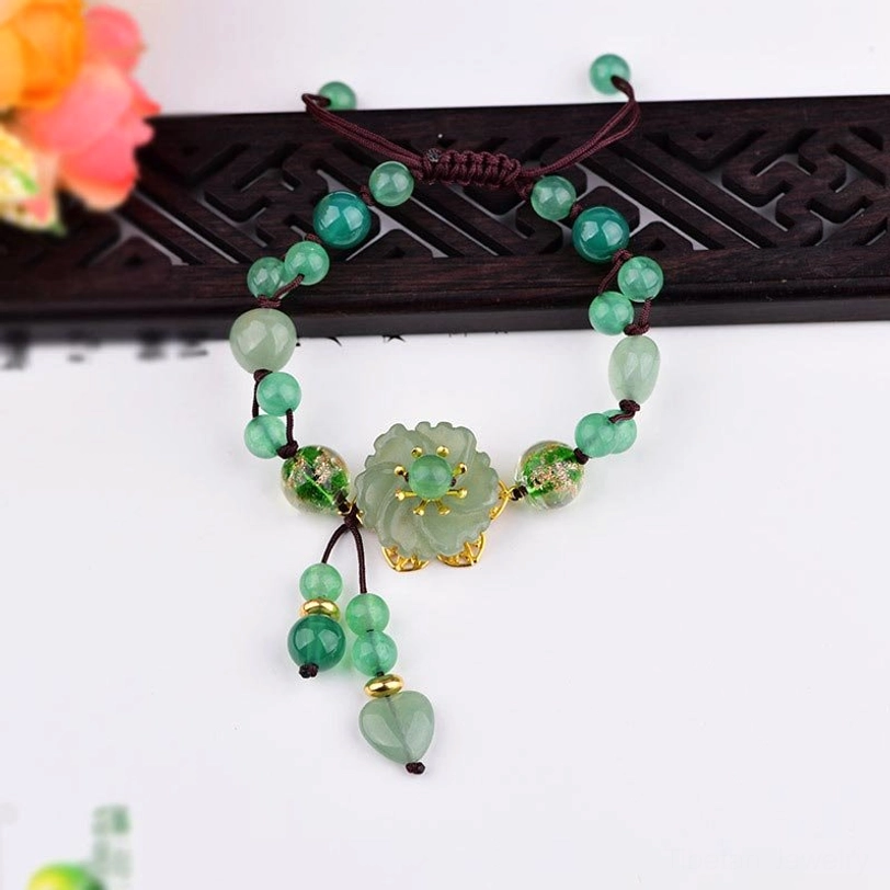 Top-down view of green jade flower dangle bracelet with braided cord and heart jade pendants on carved wooden stand