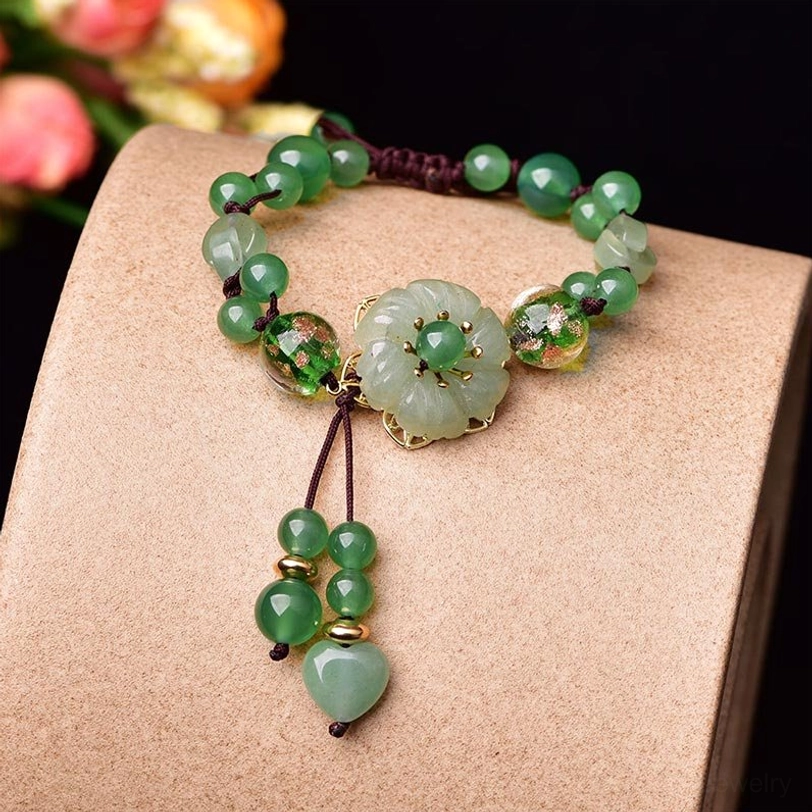 Green jade flower dangle bracelet with mixed beads displayed on light brown block showing carved flower charm and braided cord