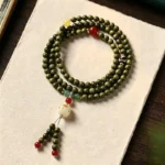 Green sandalwood cat charm mala bracelet with red accent beads, overhead shot on rice paper and wooden desk, showing multi-wrap design