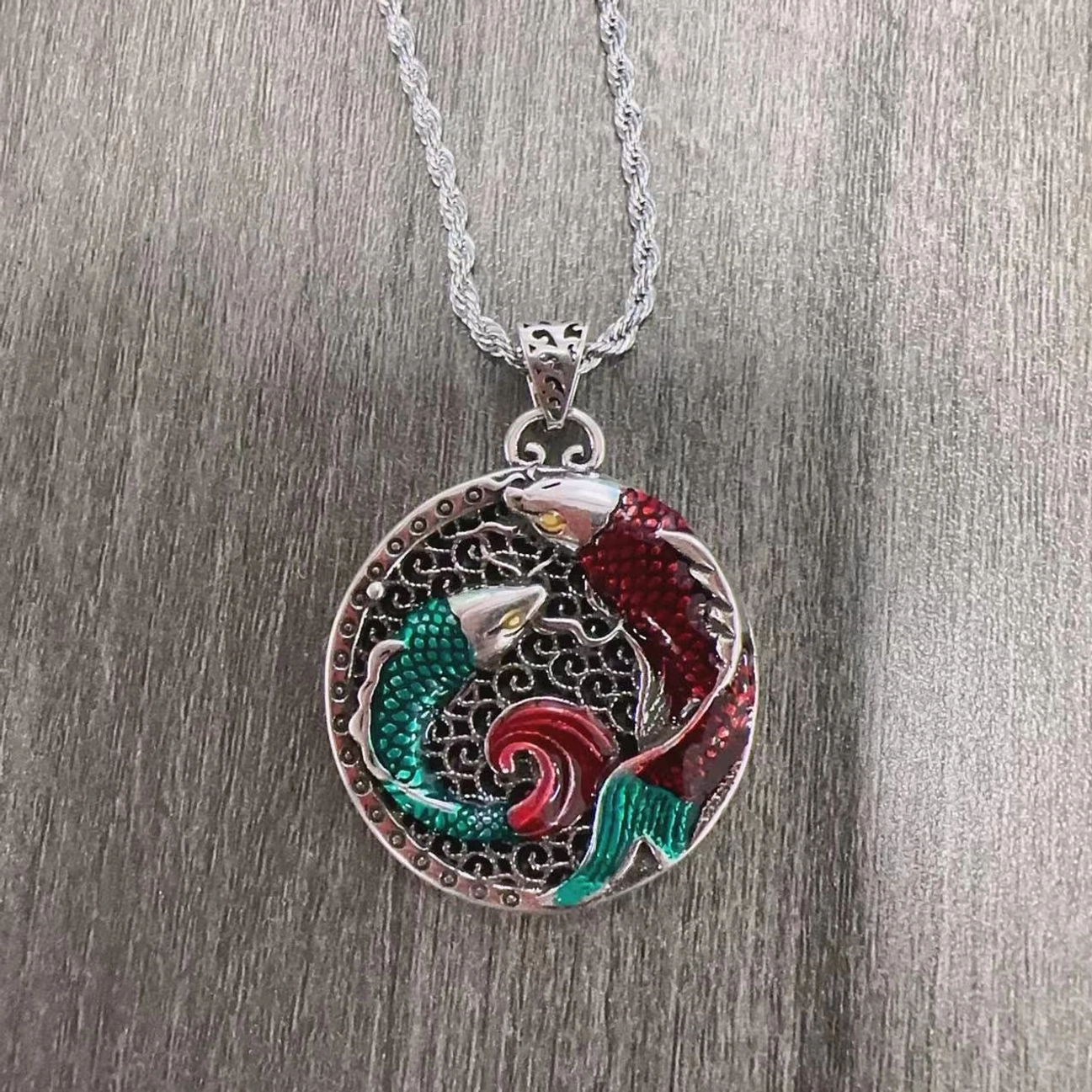 Tibetan copper koi fish round pendant with wheat-link silver chain, front close-up showing detailed hollow carved design on grey wood background