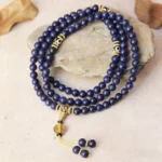 Overhead view of 108-bead lapis lazuli mala necklace with three-eyed Dzi bead and gold-tone metal spacers on stone slab with dry leaves