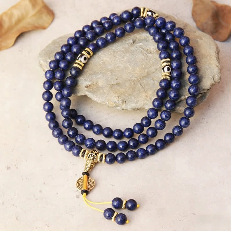 Overhead view of 108-bead lapis lazuli mala necklace with three-eyed Dzi bead and gold-tone metal spacers on stone slab with dry leaves