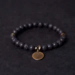 Lava rock healing bracelet with copper charm displayed flat, showing black porous beads and bronze round pendant on dark background
