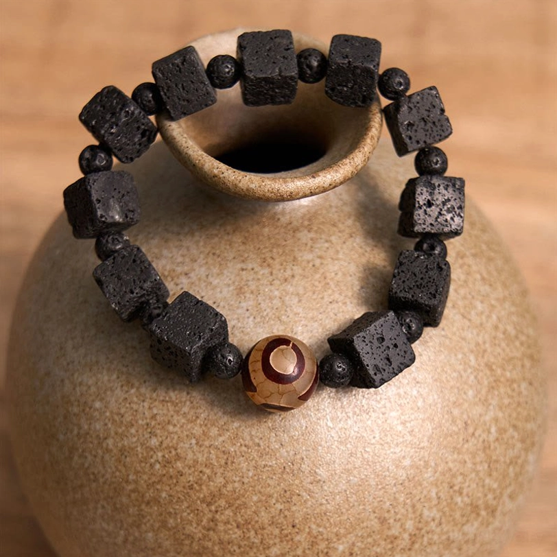 Lava Rock Dzi Bead Bracelet top-down on beige ceramic vase displaying rough dark lava cube beads, round spacer beads, and distinct patterned three-eye Dzi accent bead