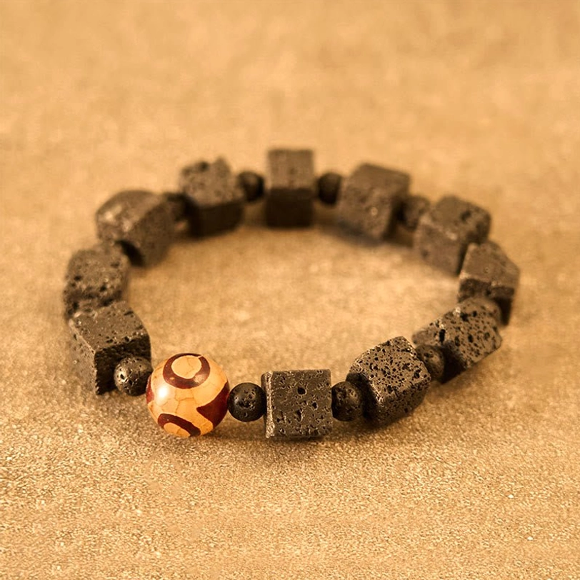 Lava Rock Dzi Bead Bracelet featuring brown-gray porous lava cube beads with orange-brown hand-painted three-eye Dzi bead and small round spacer details on tan textured background