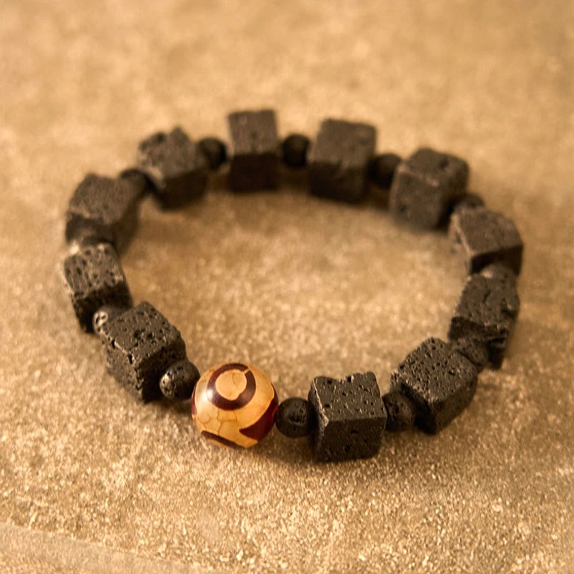 Lava Rock Dzi Bead Bracelet top-down flat lay on light brown background displaying black porous square lava beads arranged in circular pattern with central three-eye Dzi focal point