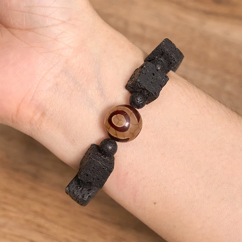 Wrist wear demonstration of Lava Rock Dzi Bead Bracelet showing black irregular lava chunks with central three-eye Dzi bead worn on human wrist against wooden background