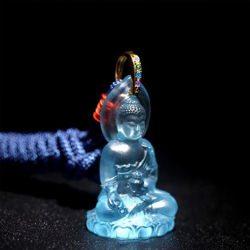 Light Blue Medicine Buddha Liuli Crystal pendant on dark background, frontal oblique view showing translucent blue crystal with gold metal bail and dark blue rope with red knot