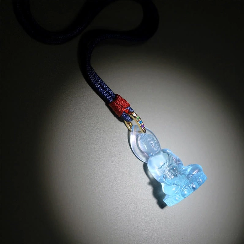 Bhaisajyaguru Buddha pendant front low-angle view on dark spotlight background, shiny translucent crystal with woven rope and red knot accent