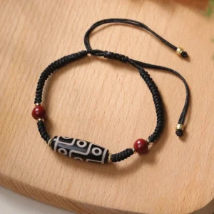 Handcrafted nine-eye dzi bead protection bracelet overhead view with black braided cord and red cinnabar accent beads on wooden surface