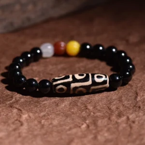 Flat lay top-down view of nine-eye dzi bead bracelet on dark brown fabric, displaying smooth black agate bead with intricate nine-eye design and multicolor stone spacers