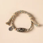 Nine-eye Dzi bead Tibetan protection bracelet with beige-blue braided rope, metal charm, and hand-painted sacred stone bead on light background