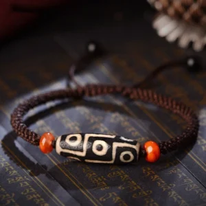 Nine-Eye Dzi Bead bracelet overhead flat lay on Tibetan scripture cloth, showcasing authentic black-and-white dzi bead with brown braided cord and orange agate spacers