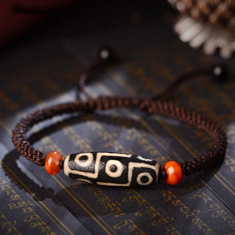 Nine-Eye Dzi Bead bracelet overhead flat lay on Tibetan scripture cloth, showcasing authentic black-and-white dzi bead with brown braided cord and orange agate spacers
