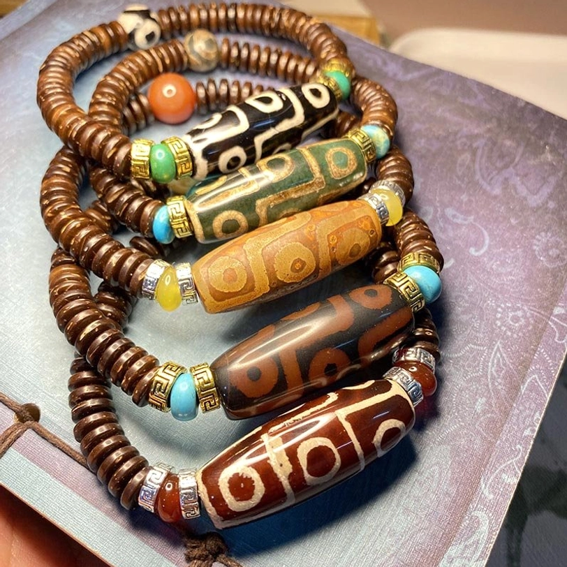 Multiple Tibetan nine-eye dzi bead bracelets stacked together showing various patterns, turquoise accents, and natural stone spacers on light purple background