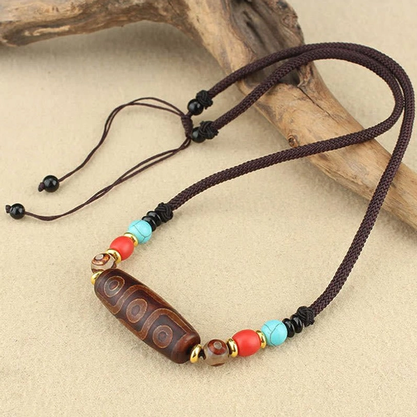 Single nine-eye dzi bead pendant necklace with adjustable brown braided cord and colorful stone beads on light wooden tabletop