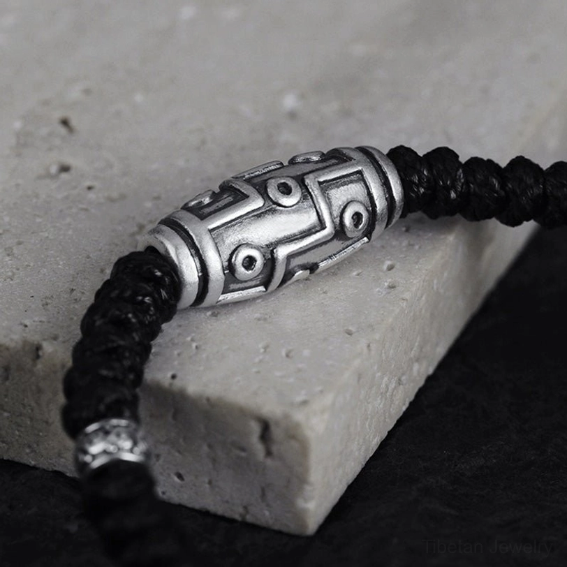 Tibetan nine-eye dzi bead bracelet low-angle side view on stone backdrop, showing silver finish and black woven cord
