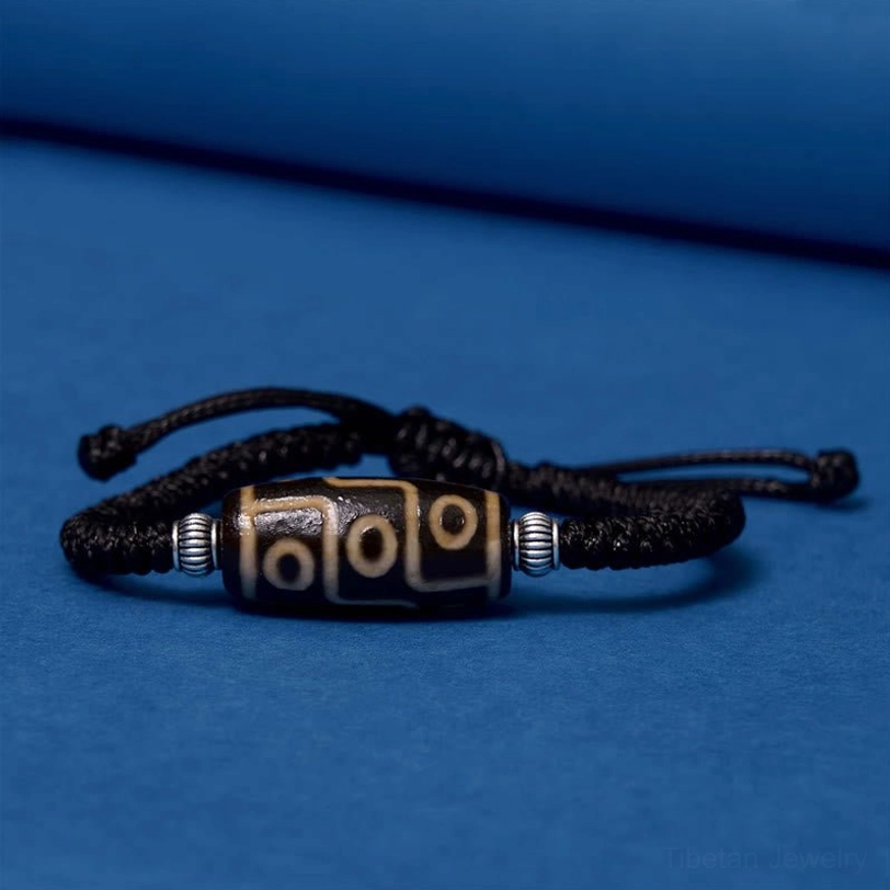 Tibetan dzi bead bracelet side profile on blue background displaying beige and black nine-eye bead with woven black cord