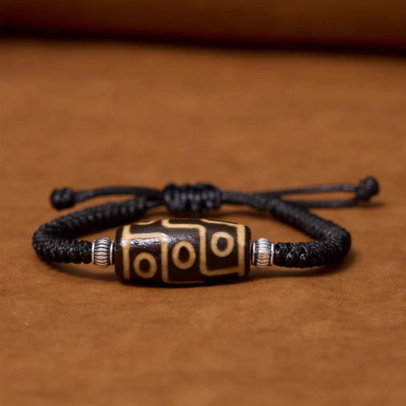 Tibetan nine-eye dzi bead bracelet flat lay overhead view on wooden surface showing black braided cord with silver spacers