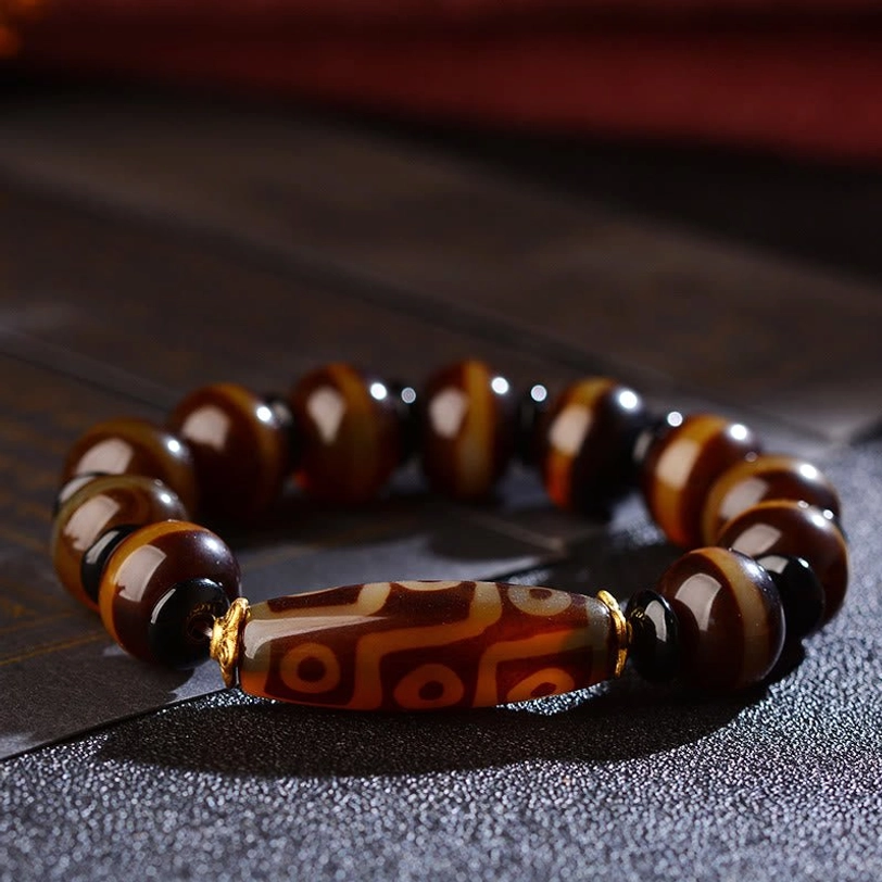 Nine-eye dzi agate bracelet side overhead flat lay on dark textured paper showing polished striped agate beads and central dzi pattern