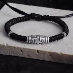 Nine-eye dzi bead sterling silver bracelet with black braided rope, top-down view showing intricate engraved pattern and silver accents
