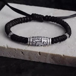 Nine-eye dzi bead sterling silver bracelet with black braided rope, top-down view showing intricate engraved pattern and silver accents