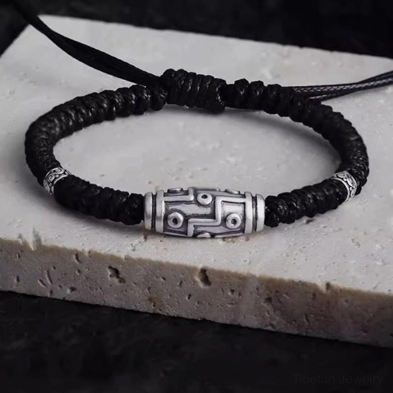 Nine-eye dzi bead sterling silver bracelet with black braided rope, top-down view showing intricate engraved pattern and silver accents