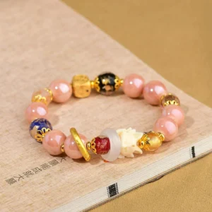 Pink luck charm bracelet with incense ash glass beads, gold hardware, blue patterned bead, red accent, and fox charm on calligraphy background