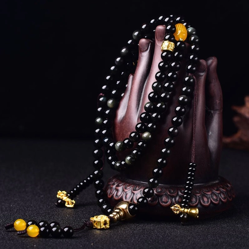 Black obsidian prayer beads mala displayed on wooden Buddha hand stand, showing polished beads and lotus tassel pendant detail
