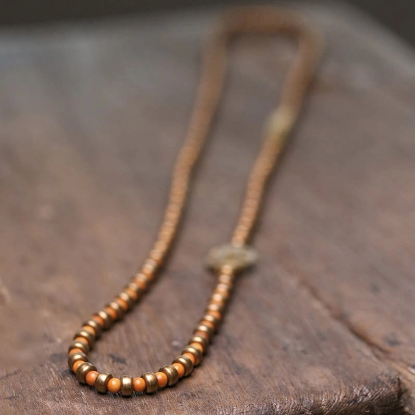 Extended olive pit and copper beaded strand displayed flat lay showing central knot accent with small brass beads on wooden background