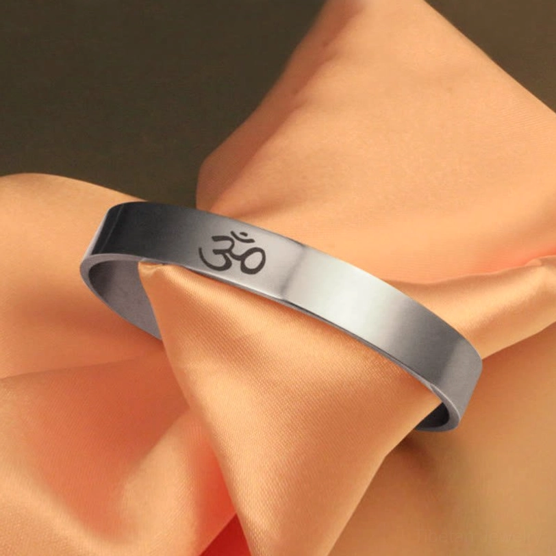 Top-down side angle of OM open cuff bracelet showing engraved symbol detail on peach satin fabric background