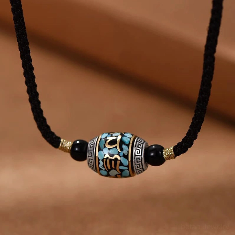 Tibetan Om Mani Padme Hum Dzi bead necklace with braided black rope chain and turquoise-inlaid pendant on brown fabric background