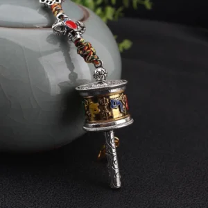 Om Mani Padme Hum prayer wheel ornament close-up showing gold-silver dual tone finish with six-word mantra carved on metal handle and ceramic pot background