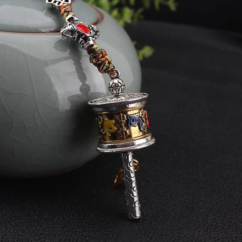 Om Mani Padme Hum prayer wheel ornament close-up showing gold-silver dual tone finish with six-word mantra carved on metal handle and ceramic pot background