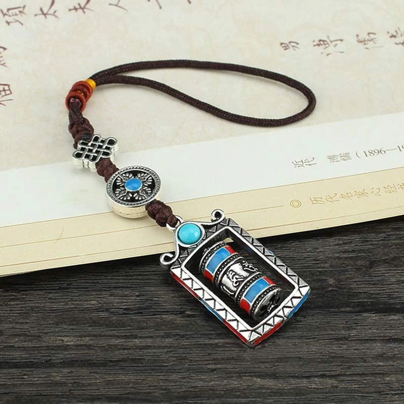 Tibetan prayer wheel pendant with Chinese knot charm, turquoise blue and silver alloy, positioned with ancient book prop on wooden surface