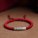Handmade Tibetan Om Mani Padme Hum sterling silver rope bracelet with dark red cord, top-down flat lay view showing adjustable knot