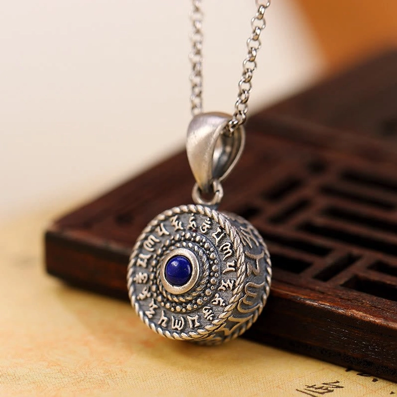 Three-quarter view of Om Mani Padme Hum Tibetan prayer pendant resting on wooden lattice and Buddhist scriptures, featuring dark matte silver and braided edge design
