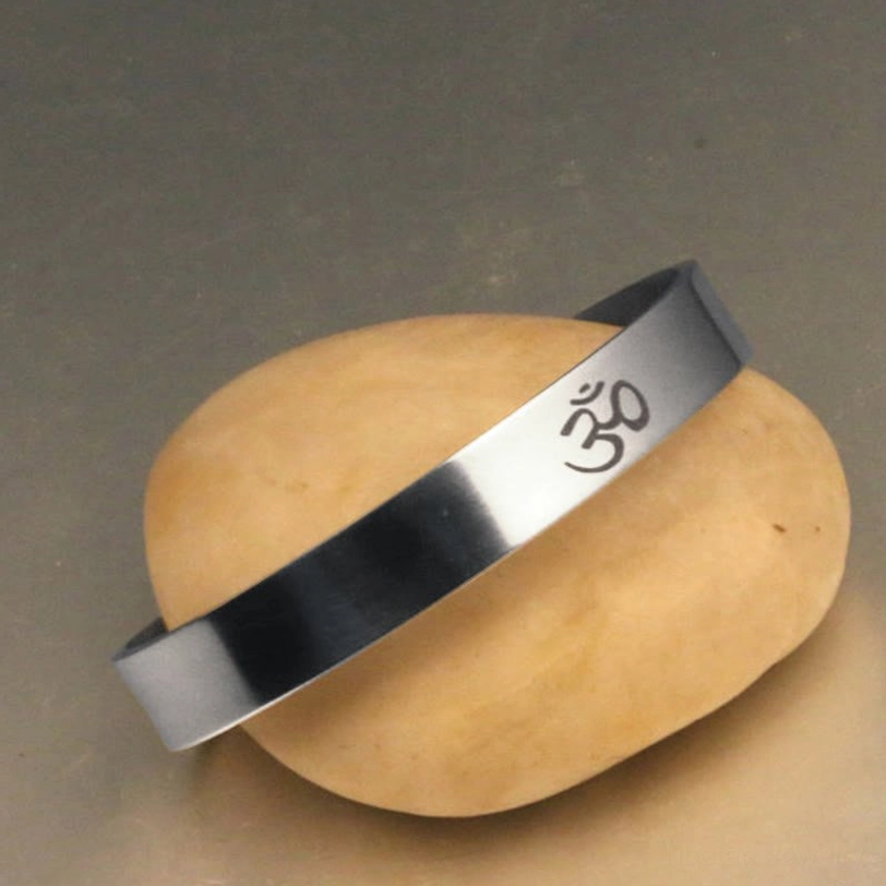 OM engraved open cuff bracelet in polished silver metal with smooth finish displayed on gray stone surface