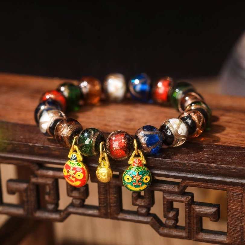 Gold Pixiu Family Charm bracelet close-up front view with two enamel pendants, one gold bell charm, and mixed bright and dark Liuli glass beads on carved wooden table edge