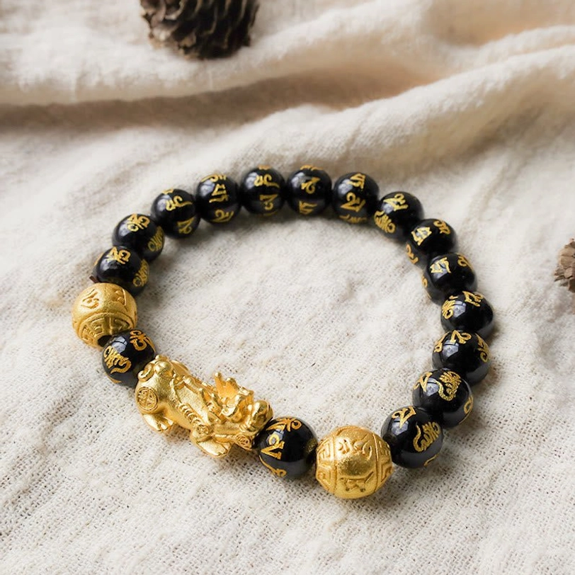 Black obsidian Pixiu prosperity bracelet with Six True Words mantra beads and golden wealth charm, flat lay view on natural fabric