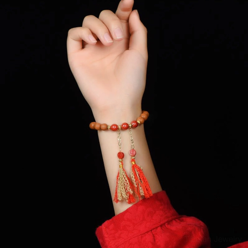 Close-up of wrist wearing peach wood cinnabar tassel bracelet with red sleeve and black background, lifestyle wear view