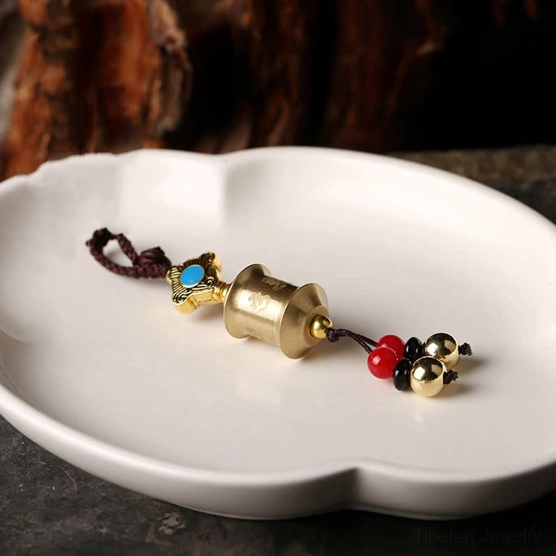 Prayer Wheel brass keychain with turquoise bead inlay and bell tassels on white ceramic plate, Buddhist meditation charm