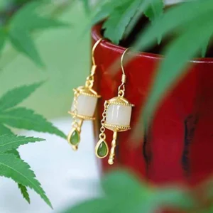 Close-up side view of prayer wheel jade drop earrings featuring sterling silver chain and decorative tassel detail next to red flower pot