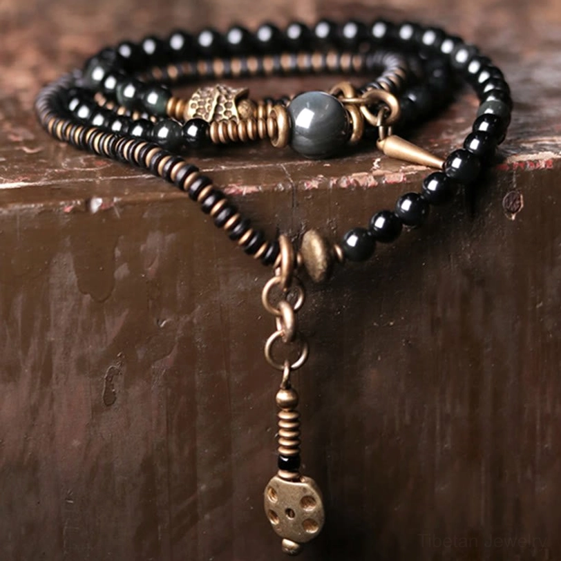 Close-up overhead view of rainbow obsidian multilayer bracelet displaying black beads with dice-shaped bronze pendant charm