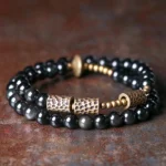 Rainbow obsidian double wrap bracelet with hammered copper accents displayed in top-down flat lay, showing polished black beads and vintage copper hardware on wooden surface