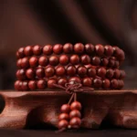 Red sandalwood 108 bead mala bracelet displayed on wooden carved base, showing front view of reddish-brown round beads with decorative tassel pendant