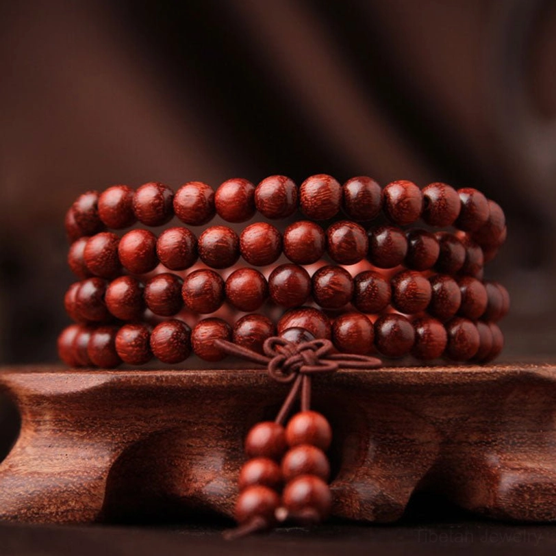 Red sandalwood 108 bead mala bracelet displayed on wooden carved base, showing front view of reddish-brown round beads with decorative tassel pendant